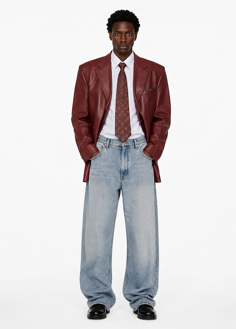 Man in a burgundy leather jacket, white shirt, patterned tie and loose light-wash jeans, standing front-on in even studio light.
