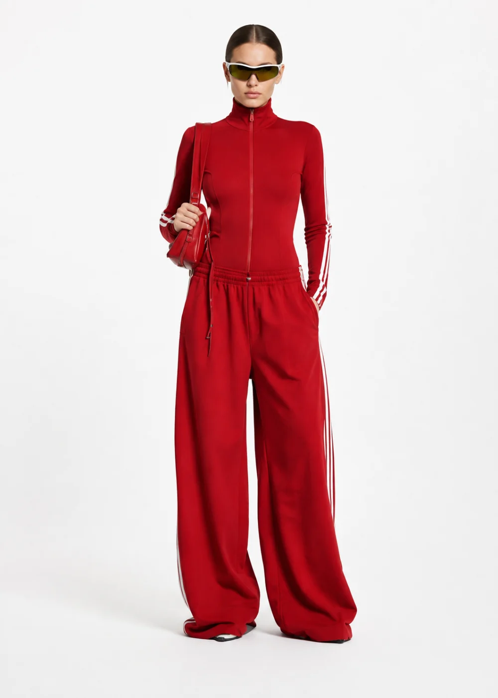 Woman in a red zip-up track jumpsuit with white side stripes, wide legs and sunglasses, carrying a small red bag, on a white background.