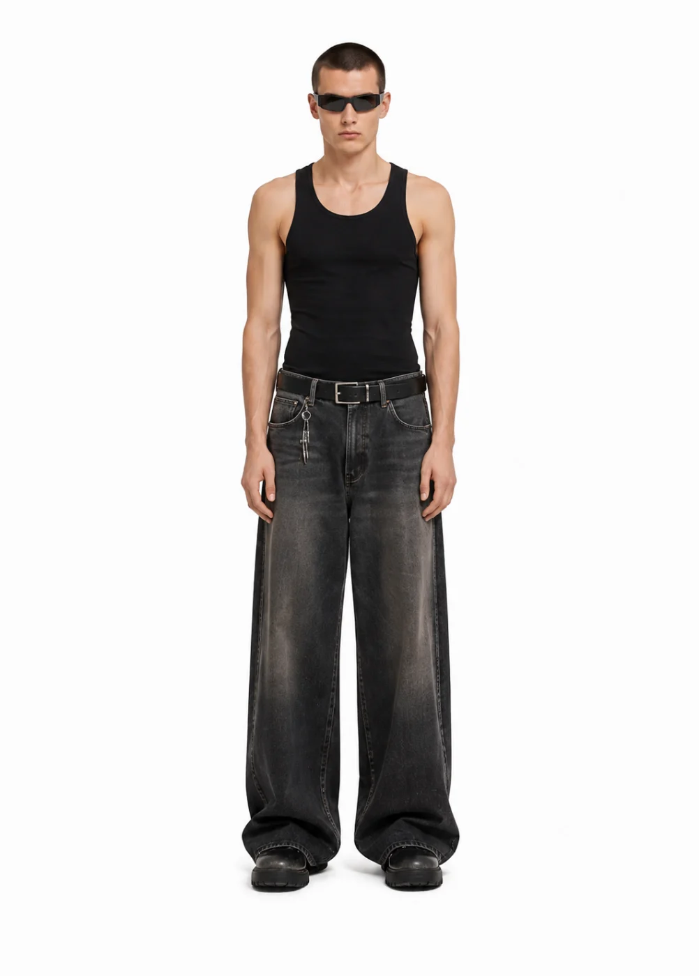 Man in a black ribbed tank top, wide faded black jeans with a leather belt, sunglasses and chunky black shoes, full length on white.