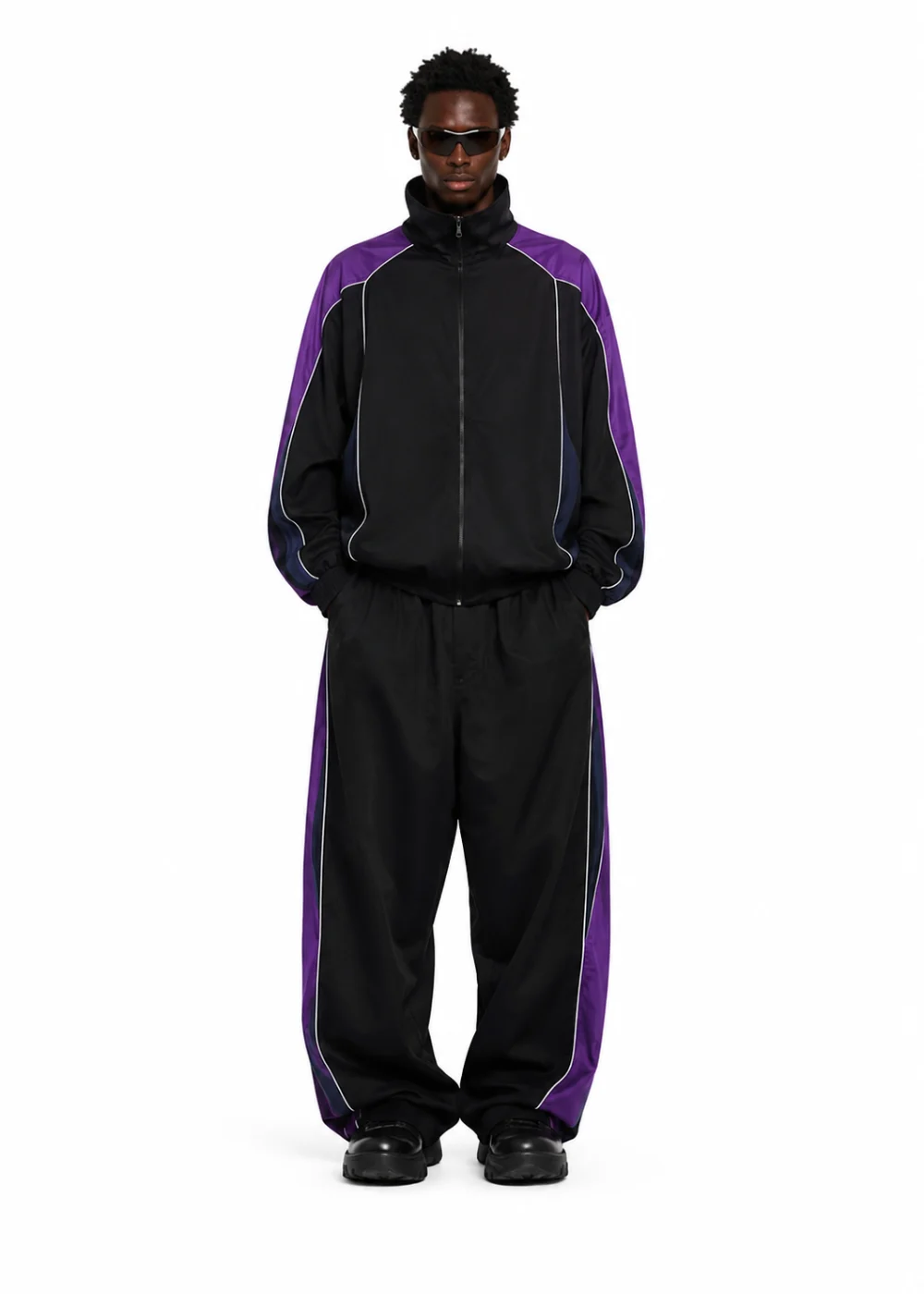 Model in a black track jacket and trousers with purple side panels and dark sunglasses, standing front-on on white.