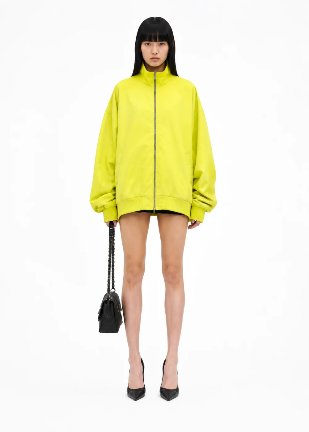 Woman in an oversized bright yellow zip-up jacket, black heels and a small quilted bag, on a white background.