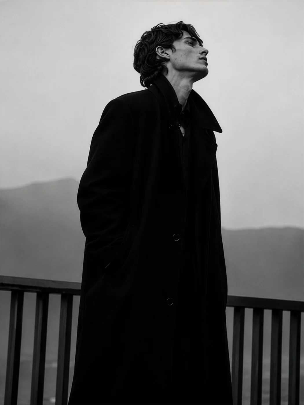 Black and white photo of a man in a long black coat on a balcony, looking up, with hills fading into the mist behind him.