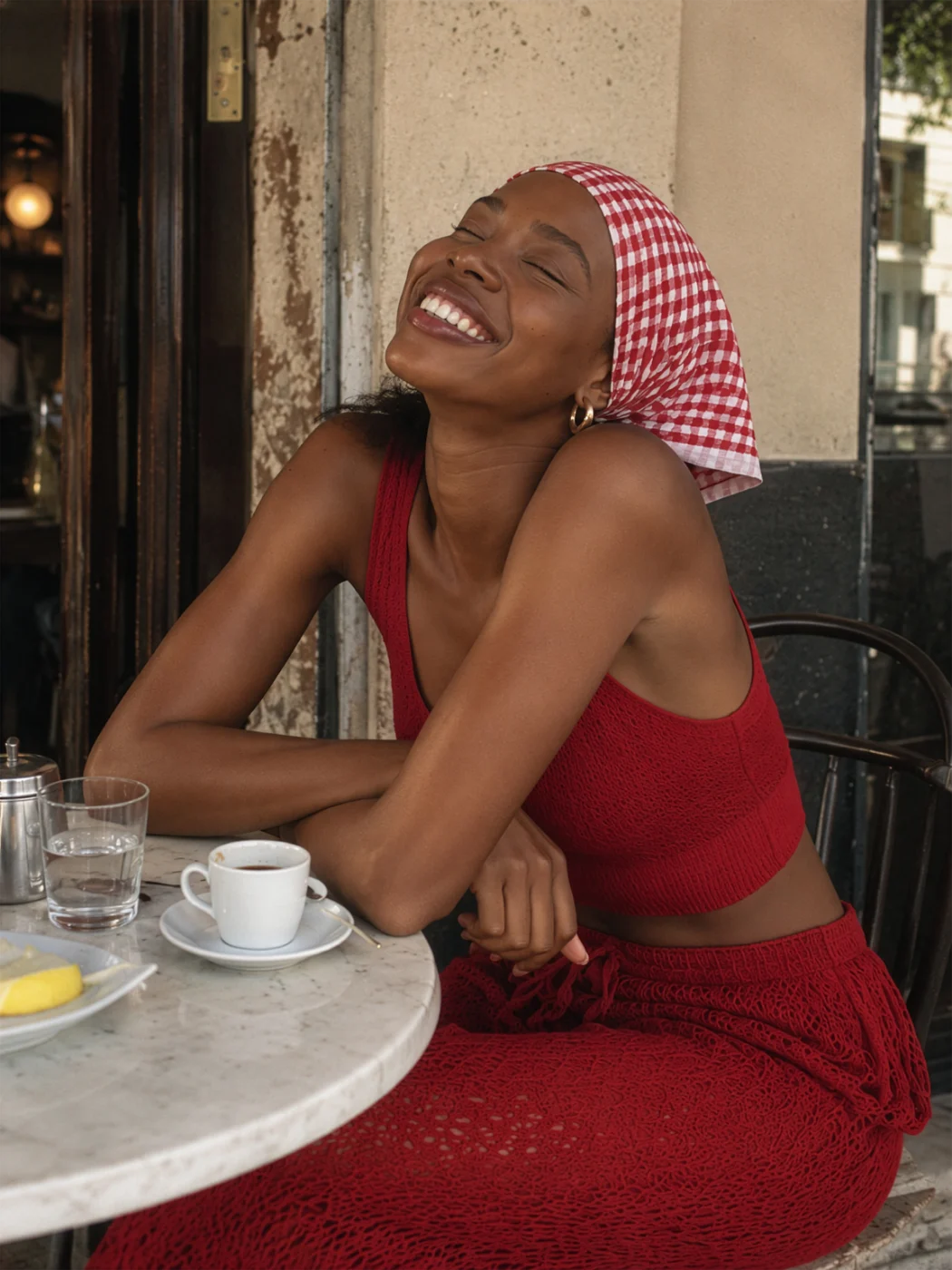 Phone-style snapshot of a smiling woman in a red knitted crop top and skirt and a gingham headscarf at a café table with coffee.