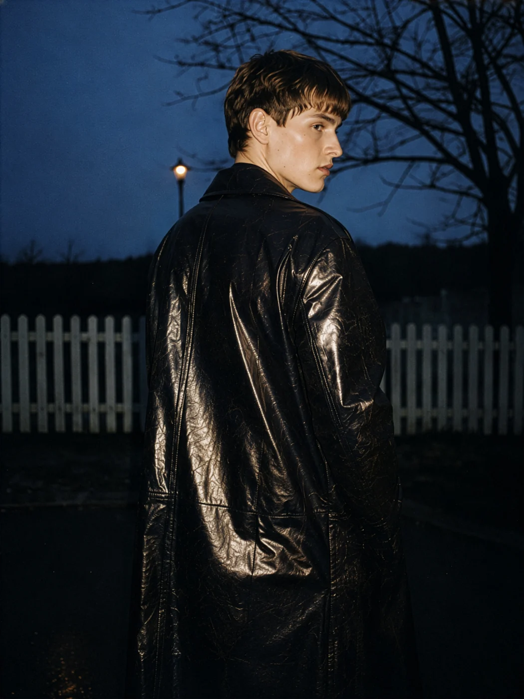 Model in a glossy black trench coat looking back over the shoulder at dusk, a white picket fence and a bare tree behind.