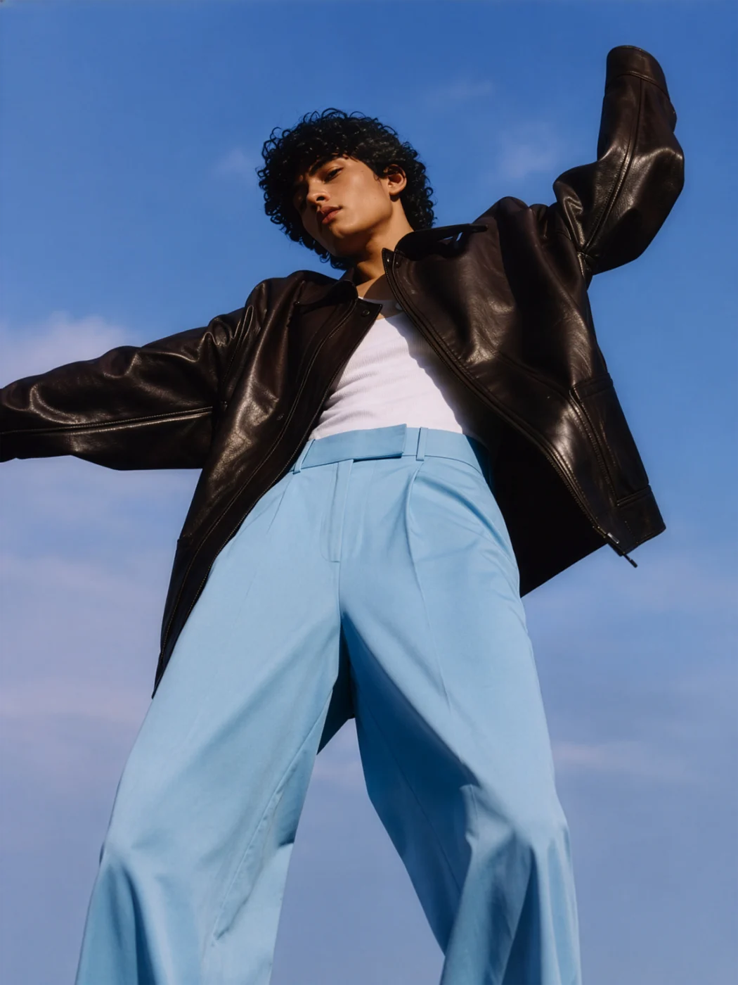 Low-angle shot of a model in an oversized brown leather jacket and pale blue wide trousers, arms out against a bright blue sky.