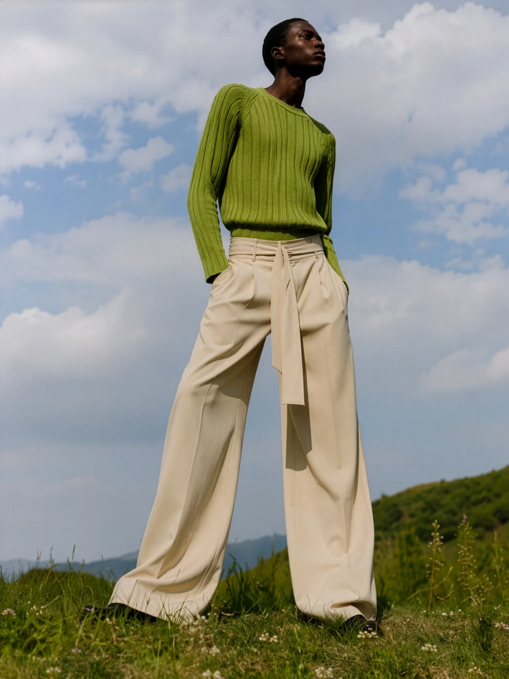 Model in a green ribbed sweater and wide cream trousers standing on a grassy hill under a cloudy sky.