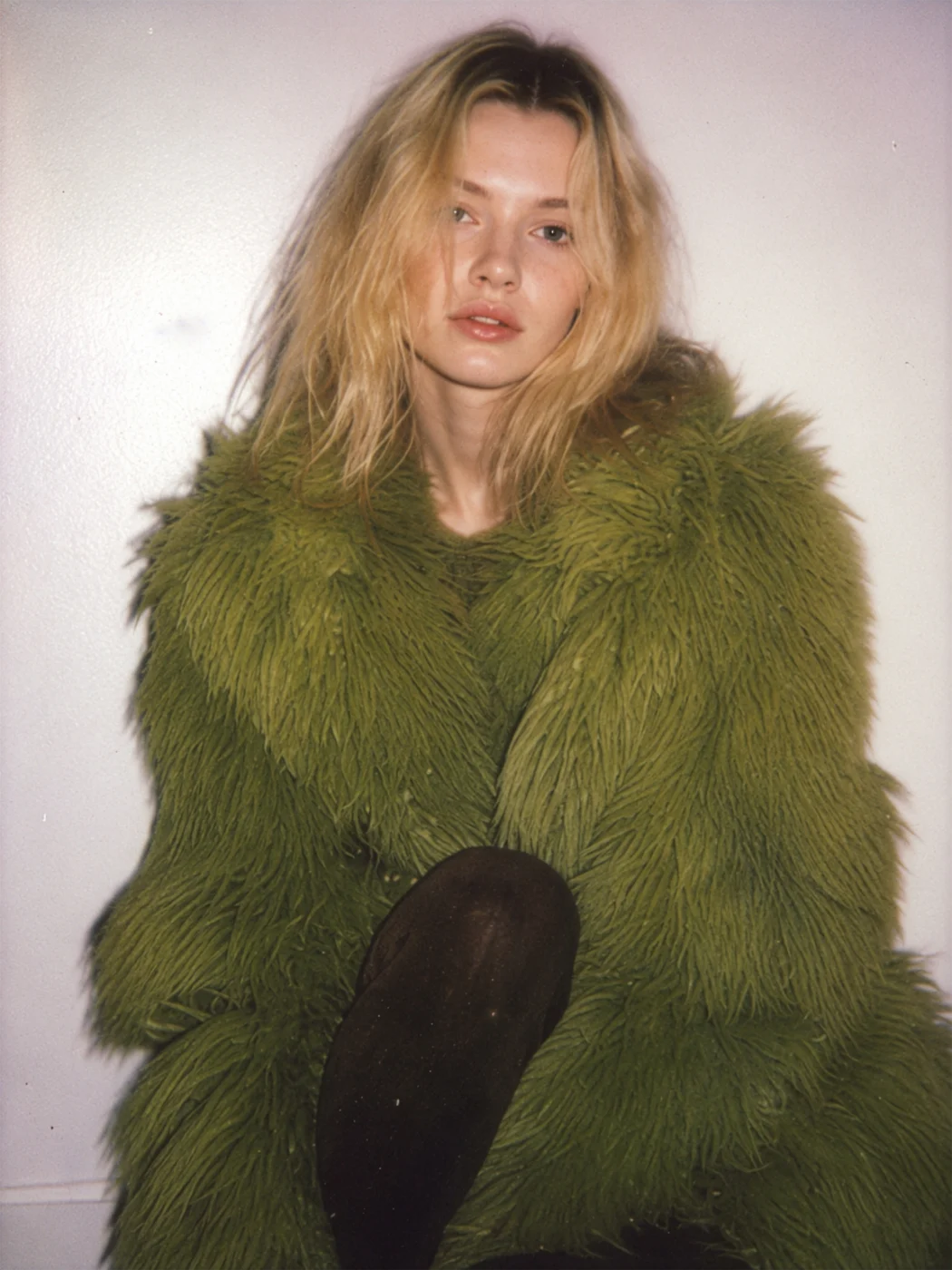 Instant-film style photo of a blonde model sitting in a fluffy green faux fur coat against a plain wall, lit by a soft flash.