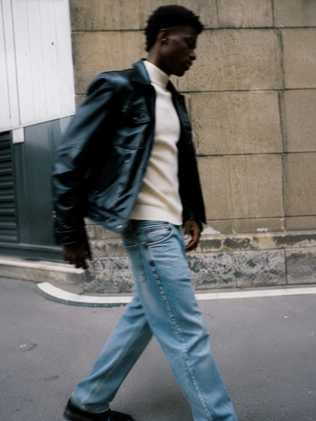 Man caught mid-stride past a stone building in a black leather jacket, cream shirt and light blue jeans.