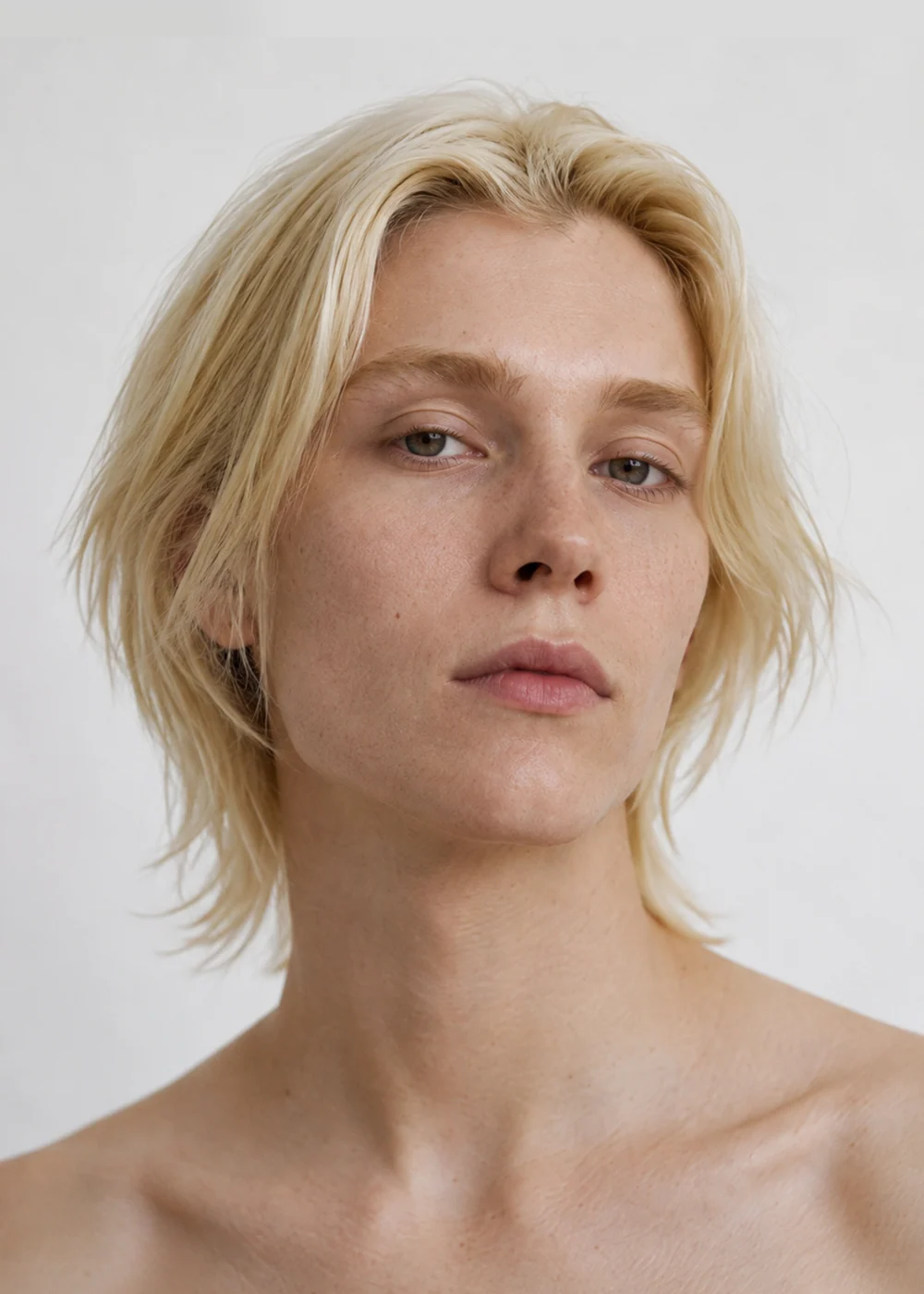 Portrait of a non-binary model with chin-length blonde hair and blue eyes, relaxed neutral expression, white background.