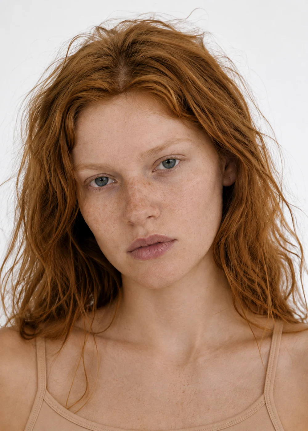 Portrait of a young woman with wavy copper-red hair, freckles and blue eyes, wearing a beige camisole.