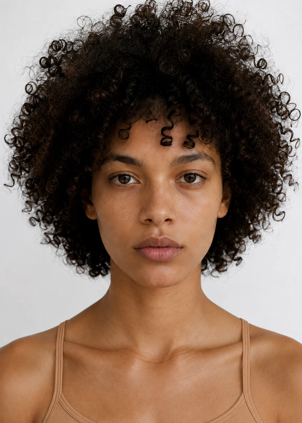 Portrait of a young woman with voluminous dark curls and hazel eyes, wearing a tan camisole.