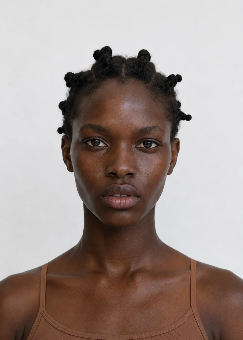 Portrait of a young woman with bantu knots and dark brown skin, brown camisole, neutral expression on a light grey background.