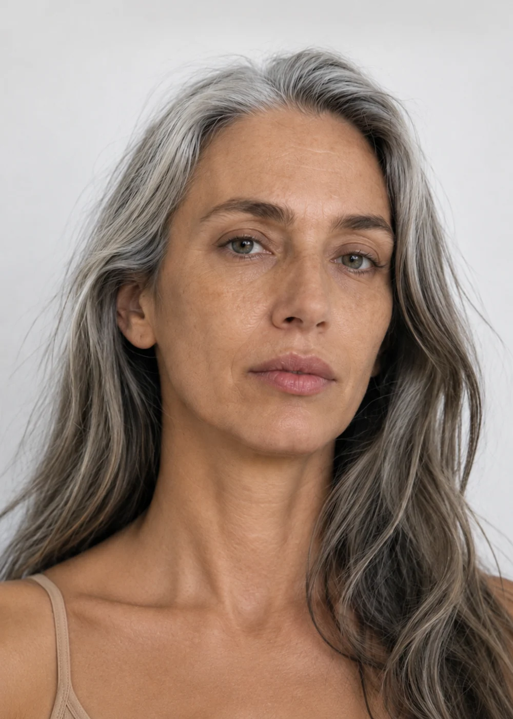 Casting portrait of a woman in her fifties with long grey hair and a beige camisole, soft light on a white background.