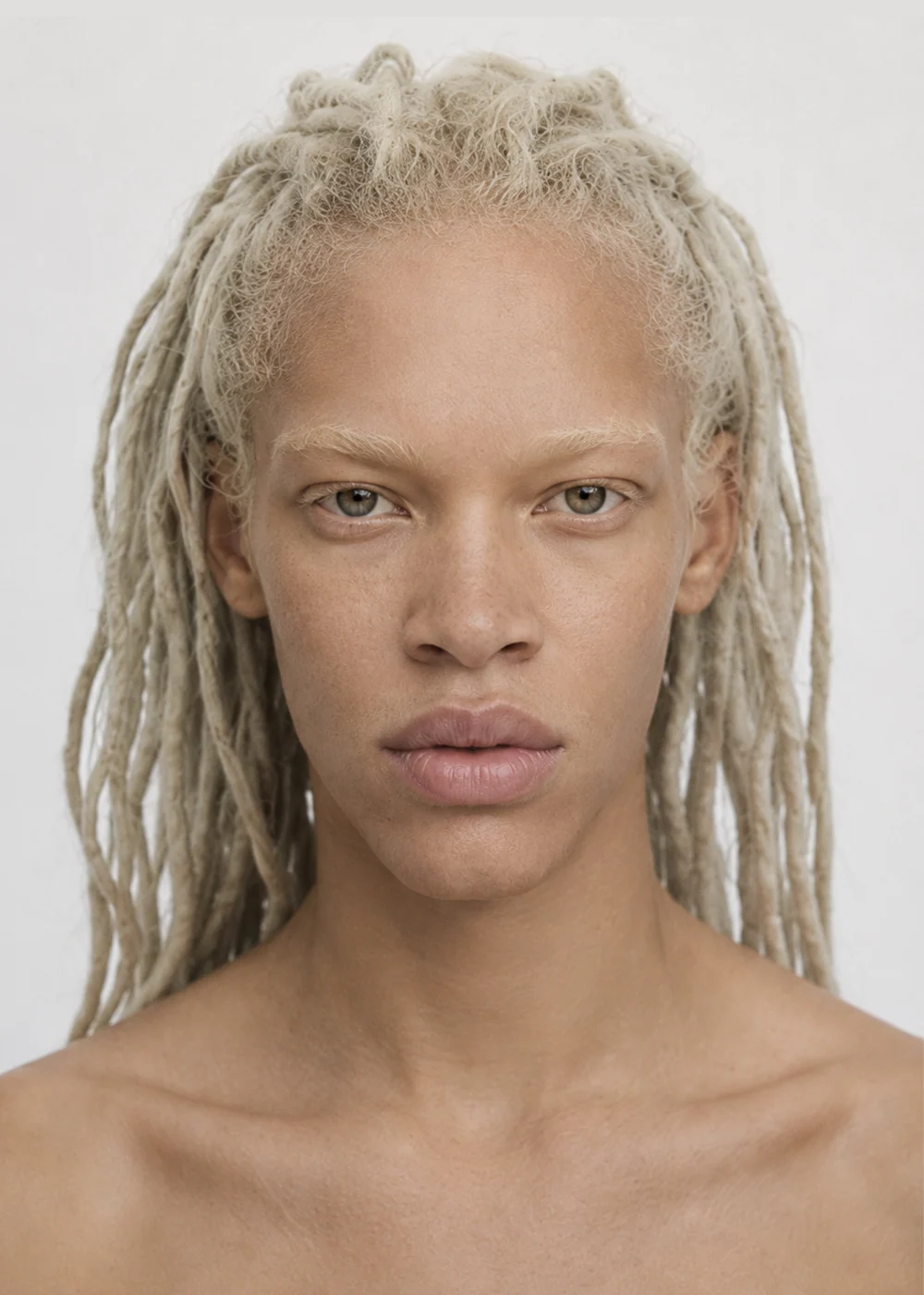 Portrait of a young model with pale blonde locs, light eyebrows and grey eyes, neutral expression against a white wall.